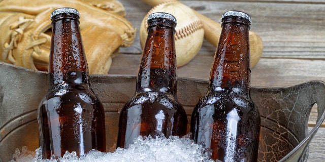 Close up of three brown cold bottled beers, crushed ice in metal container, and baseball equipment in background on rustic wood