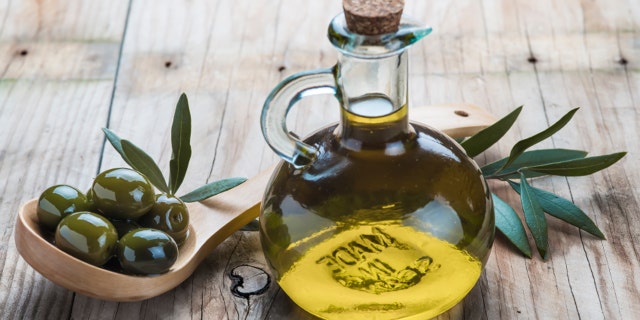 A glass bottle of olive oil and a wooden spoon with olives on a table