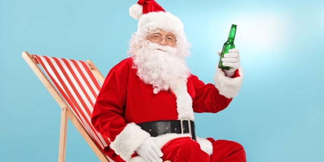 Santa Claus holding a bottle of beer seated on a sun lounger and looking at the camera with blue sky in the background