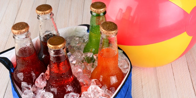 Closeup of a cooler with soda bottles and ice on a rustic wood table with a beachball in the background.
