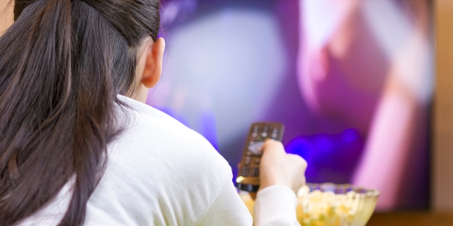 Teenager girl with remote control laying down and watching tv eating popcorn.