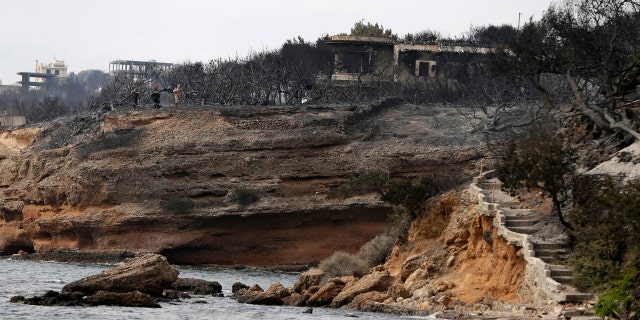 Firefighters stand on a cliff top where burned trees hug the coastline in Mati east of Athens.