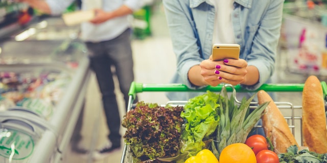 Couple in the supermarket. Cropped image of girl leaning on shopping cart, using a mobile phone and smiling, in the background her boyfriend is choosing food