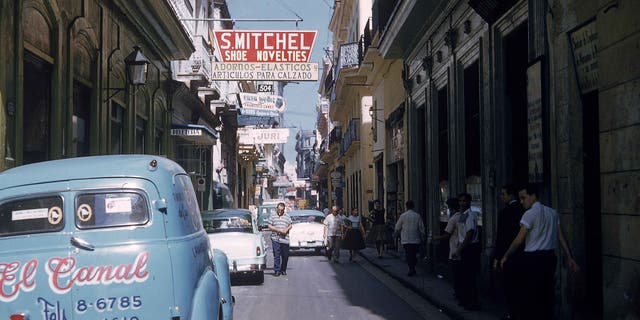 Cars are parked along a narrow street as pedestrians walk in the shade in Havana, Cuba, 1950s.