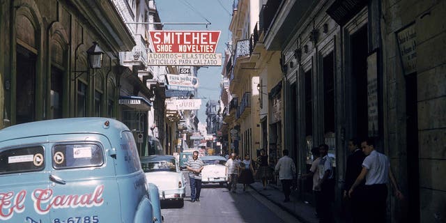 Cars are parked along a narrow street as pedestrians walk in the shade in Havana, Cuba, 1950s.