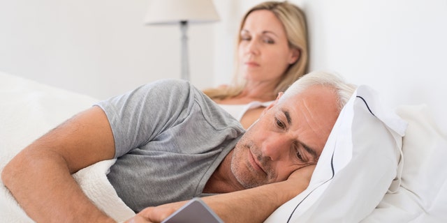 Jealous woman spying her husband mobile phone while he is reading a message. Senior couple in bed while wife is angry as husband using smartphone. Senior husband ignoring wife and texting on smartphone.