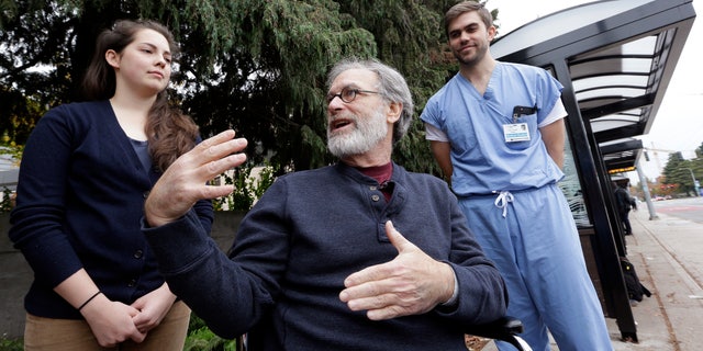 Stephen DeMont, center, sits in a wheelchair at the bus stop where he says that Zach Forcade, right, and Madeline Dahl saved his life days earlier in front of the University of Washington Medical Center, Wednesday, Oct. 19, 2016, in Seattle. When DeMont collapsed at a bus stop in front of the UW Medical Center on his morning commute, Dahl was one of 41 people within a 330-yard radius who happened to have a cell phone app alerting them to the emergency. Forcade, a medical student, witnessed the collapse and rushed over to begin chest compressions, as within moments Dahl, a cardiac nurse just getting off her shift in the hospital, was alerted by her phone and sprinted down the sidewalk, assisting until paramedics arrived. (AP Photo/Elaine Thompson)