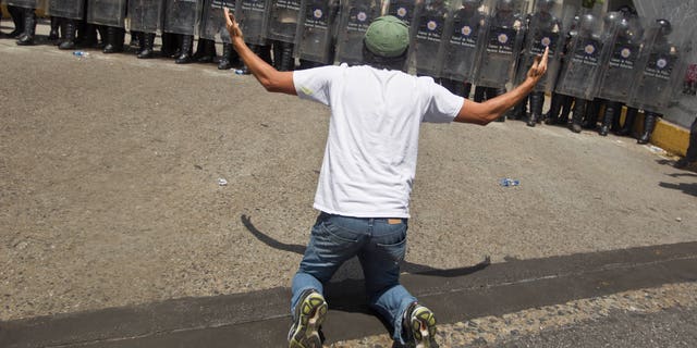 An anti-government demonsrator kneels in front of a formation of Bolivarian National Police in riot gear, during clashes at the Central University of Venezuela, UCV, in Caracas, Venezuela, Thursday, March 20, 2014. Thursday dawned with two more opposition politicians, San Cristobal Mayor Daniel Ceballos and San Diego Mayor Enzo Scarano, behind bars. Police used tear gas and water cannons to disperse a student-called protest of several thousand people in Caracas, some of those demonstrating against the arrests of the mayors. (AP Photo/Esteban Felix)