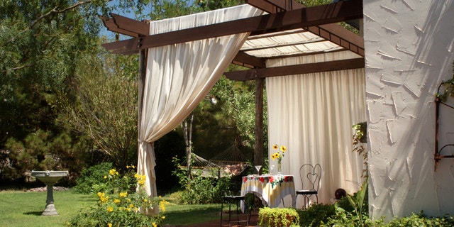 A brick patio with wooden patio posts and a cover of canvas curtains. The back ground has green grass, a birdbath and hammock.