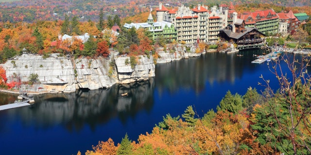 The blue sky and the Mohonk Mountain House resort reflected in Mohonk Lake and surrounded by sandstone cliffs and colorful Autumn trees in the Shawangunk Mountains of New York