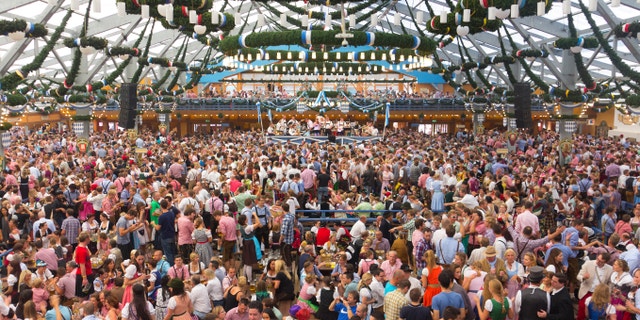 At the oktoberfest Munich. Hundreds or maybe thousands of poeple are inside one of the big beertents, sitting or standing on their benches, talking, drinking and having fun. Many of them dressed in dirndls or leather pants. In front is the band playing drinking songs.