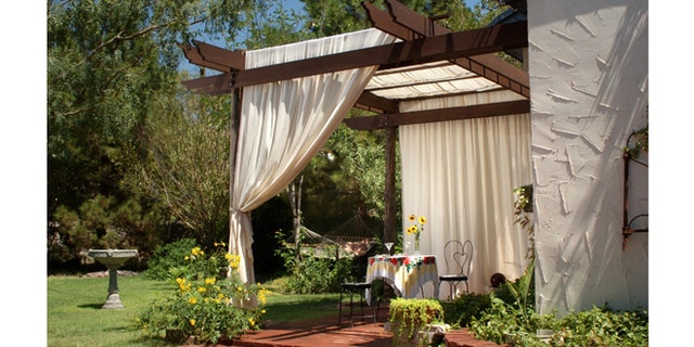A brick patio with wooden patio posts and a cover of canvas curtains. The back ground has green grass, a birdbath and hammock.