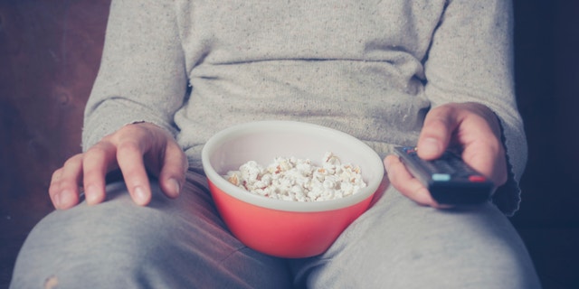 Young man is sitting on a sofa and eating popcorn while watching television