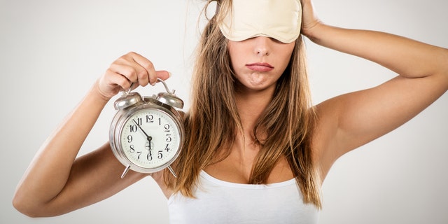 Beautiful young woman with sleeping mask holding alarm clock. She is tired and lazy in morning.