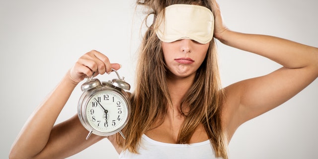 Beautiful young woman with sleeping mask holding alarm clock. She is tired and lazy in morning.