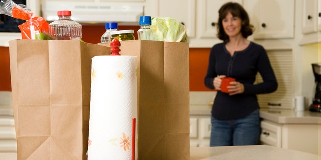 Woman in the kitching with cup of coffee, she is looking at the grocery bags to be unloaded that are on the counter.