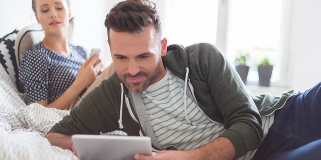 Man in casual outfit lying in the bed at home and using a digital tablet. His girlfriend in the background.