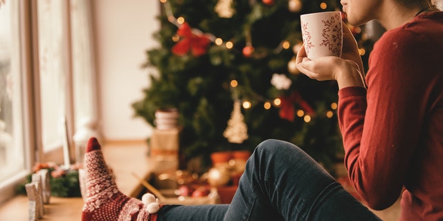 Young woman drinking tea by the Christmas tree, looking through window.