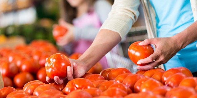 A woman is handling fresh tomatoes at a supermarket or local grocery store. Her daughter is also choosing vegetables in the background, adding depth and context to the image.