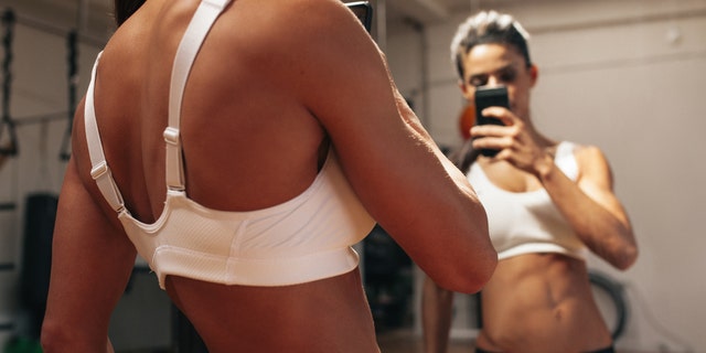 Sporty young woman taking a picture of herself in a mirror. Fitness model taking a selfie in front of a mirror in gym.