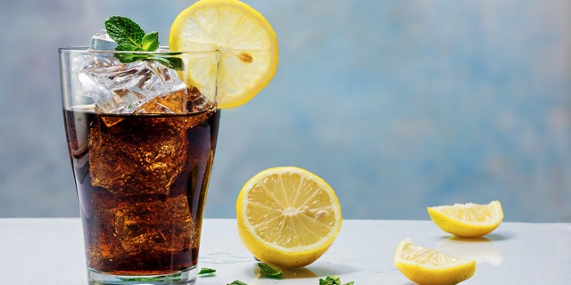 glass of cola or ice tea with ice cubes, lemon slice and peppermint garnish, against a blue wall
