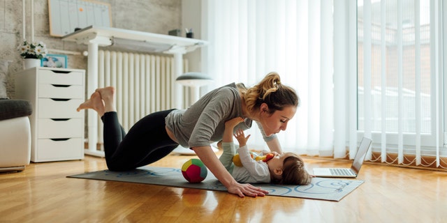 Shot of an attractive young woman with her baby girl while doing exercise. Woman working pushups and baby lying on exercise mat. They exercise in a living room. Woman watching exercise on laptop.
