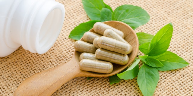Herb capsule spilling out of a white bottle in wooden spoon with green leaf on sack background.