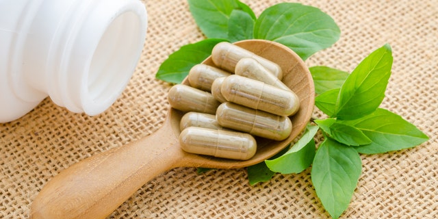 Herb capsule spilling out of a white bottle in wooden spoon with green leaf on sack background.
