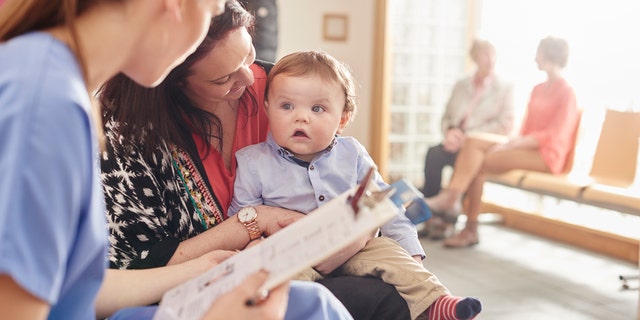 a female nurse sits next to a woman and her baby son in the waiting room of a clinic and casually chats to them . In the background two patients sit and chat to a senior doctor .