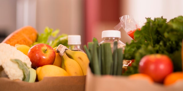 Bags full of groceries in paper sacks on a counter in a home kitchen. Food items include: apples, bread, lettuce, bananas, cauliflower and a variety of other vegetables and food items. Domestic kitchen can be seen in background.