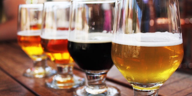 Flight of four beers lined up on a wooden table, with selective focus on the first beer. Photo taken outdoors with natural lighting; beers range in color from light to very dark. Background out of focus.