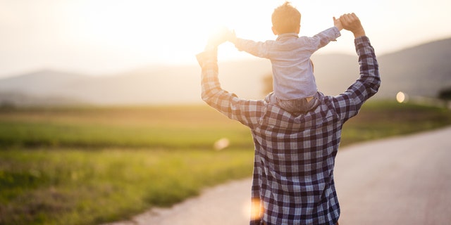 Father carries his baby boy on his shoulders while they are walking and having fun on the country road