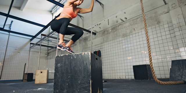 Fit young woman box jumping at a crossfit style gym. Female athlete is performing box jumps at gym.