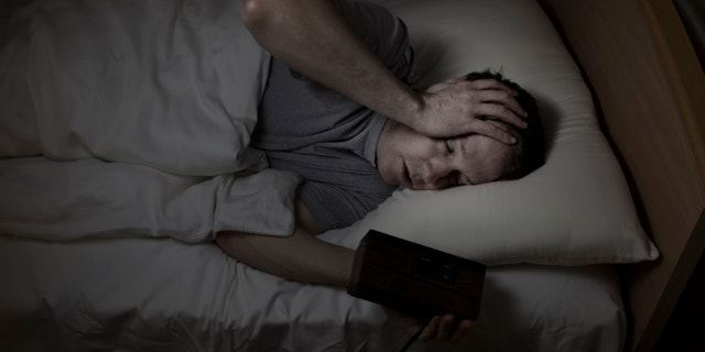 Mature man, eyes open with one hand holding clock and the other his own forehead, cannot sleep at night from insomnia