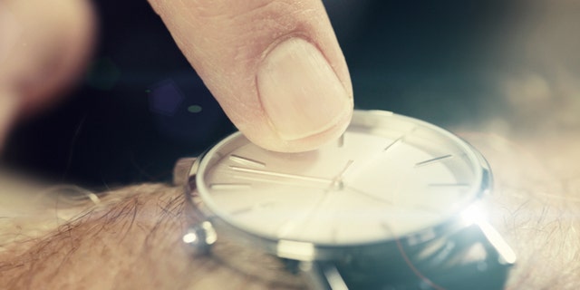 A man's finger taps impatiently on the dial of a watch, indicating time pressure or lateness.