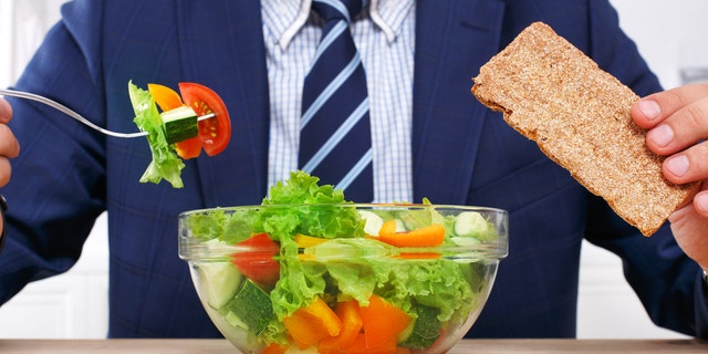 Man in suit unrecognizable torso eating healthy business lunch in modern office interior. Businessman at working place with vegetable salad in bowl, diet and right nutrition concept.