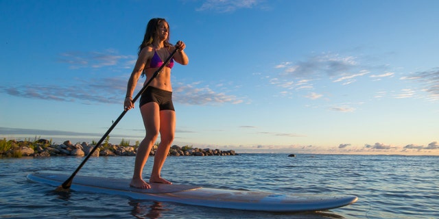A lone woman paddles her stand up paddle board through the waves as the sun rises.