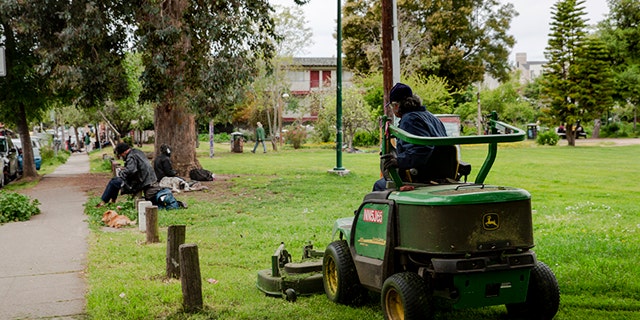 A worker mows the lawn in People's Park while a homeless man sits on the grass.