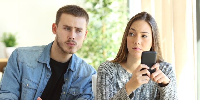 Jealous boyfriend spying his girlfriend watching her phone while she is looking him upset (Credit: AntonioGuillem, iStock)