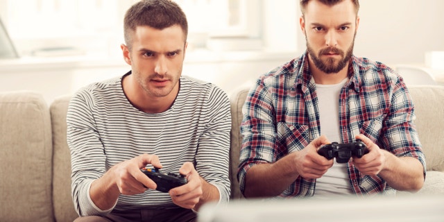 Focused on game. Two concentrated young men playing video games while sitting on sofa. (iStock)