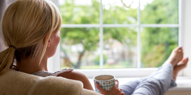 A young woman sits with a hot drink and her feet up, watching out of the window from the comfort of her own home. She is taking a break from her chores and busy day to day routine to gaze into her garden and wonder.