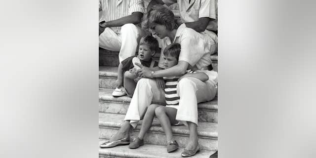Princess Diana puts on shoes for little Prince Harry as her other son Prince William squints from the bright sun during their vacation with the Spanish royal family at Marivent Palace in Palma De Mallorca.
