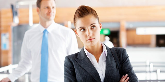 Frustrated businesswoman waiting with male executive standing in background at airport. Horizontal Shot.