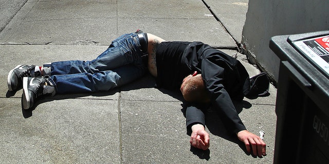 April 26, 2018: A man lies on the sidewalk beside a recyclable trash bin in San Francisco. The city's voters will decide in November 2018 whether to tax large businesses to pay for homeless and housing services in a city struggling with income inequality. Supporters collected enough signatures to get the measure on the ballot.