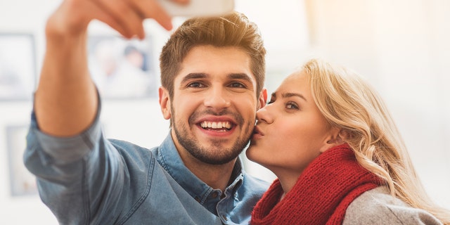 Cheerful loving couple is making selfie on smartphone. Man is looking at camera and smiling. Woman is kissing him with love