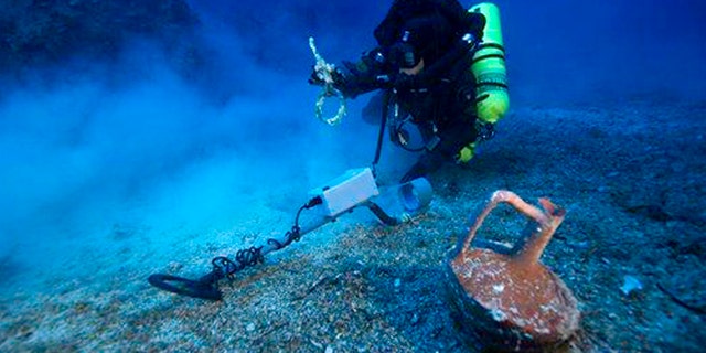 In this undated photo provided by Argo via the Greek Culture Ministry, a diver with a metal detector holds a copper ship's fitting next to a vase at the site of the Antikythera wreck off the island of Antikythera in southern Greece.