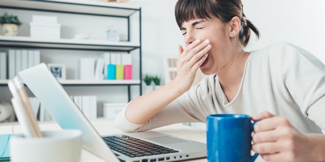 Tired sleepy woman yawning, working at office desk and holding a cup of coffee, overwork and sleep deprivation concept