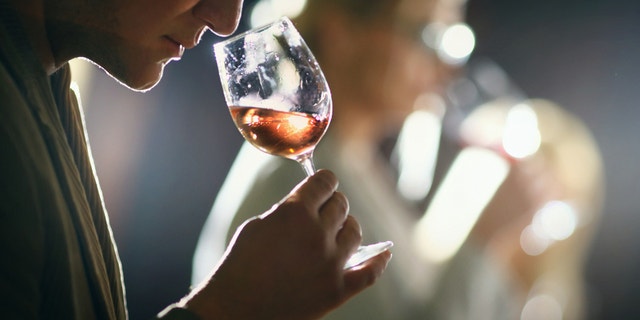 Group of unrecognizable caucasian adults tasting wine in wine cellar. Comparing appearance, smell, aroma,taste,aftertaste. The man in focus is holding a glass of rose wine and smelling the bouquet. There are two more people in background,blurry.