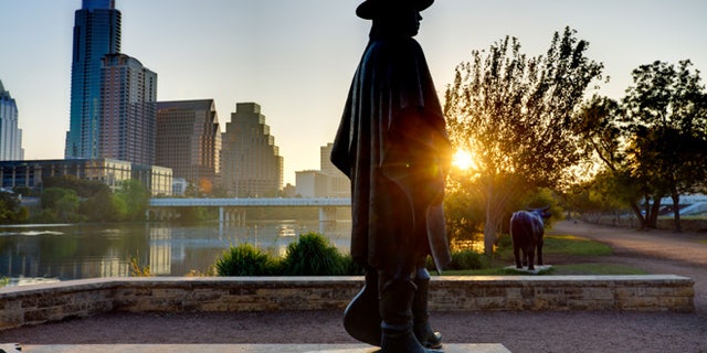 The statue of Stevie Ray Vaughan stands on the shores of Lady Bird Lake, with the skyline of Austin behind him.