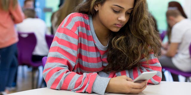 Female student sitting on her own at school. She has a smartphone in her hand and a stressed expression on her face.